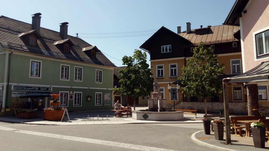 Blick auf das Kriegerdenkmal in Bad Mitterndorf mit dem Sportgeschäft Mörth und dem Restaurant S’Platzl. Im Hintergrund strahlt ein blauer, wolkenloser Himmel. | © Johanna Provatopoulos