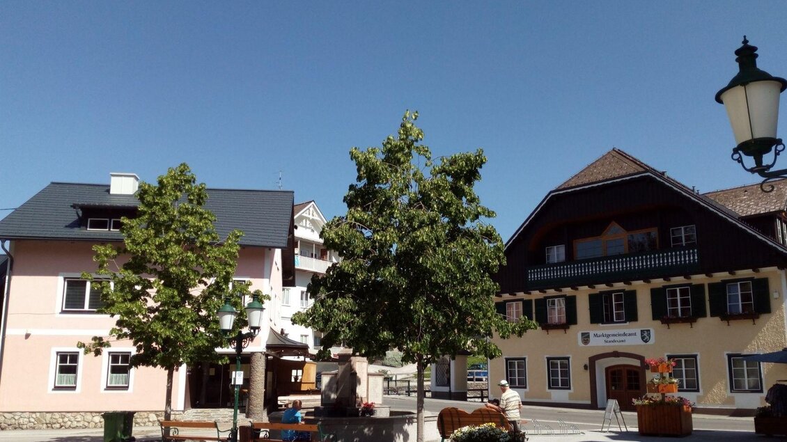 Hinteransicht mit wunderschönem Blick auf das Gemeindeamt der Marktgemeinde Bad Mitterndorf und das S’Platzl. Zwei kleine Bäume und Bänke säumen den Brunnen. Darüber strahlt ein strahlend blauer, wolkenloser Himmel. | © Johanna Provatopoulos