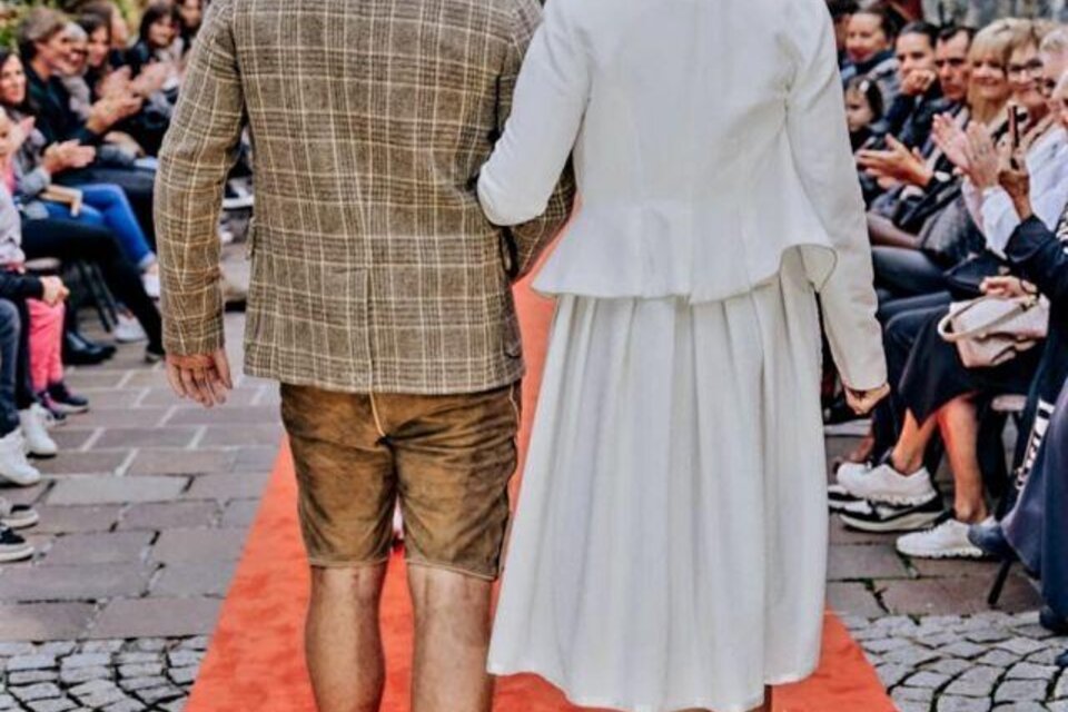 Ein Paar geht auf einem roten Teppich entlang einer Menschenmenge. Die Frau trägt ein elegantes weißes Outfit, der Mann eine schicke Kombination aus Blazer und Shorts. | © Stefanie Pirstinger Photography