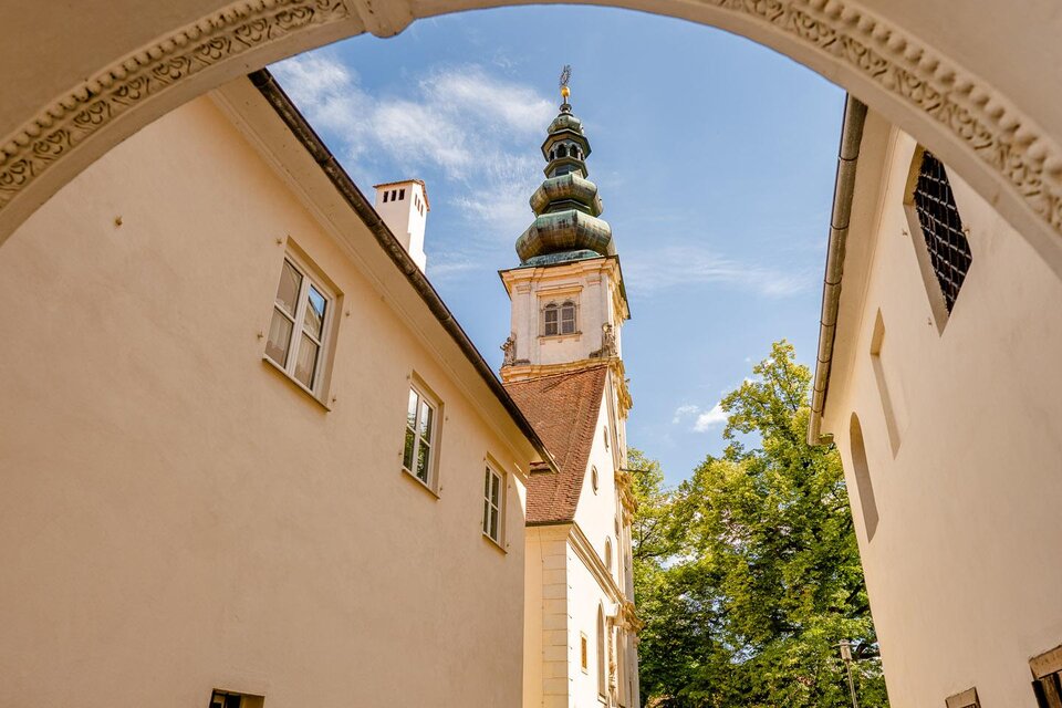Ein malerischer Durchgang führt zu einem historischen Gebäude mit einem hohen Turm. Umgeben von grünen Bäumen und einer schönen Umgebung spazieren Menschen entlang des Weges. | © Wolfgang Spekner
