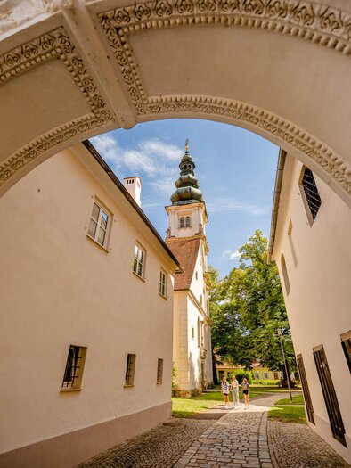 Ein malerischer Durchgang führt zu einem historischen Gebäude mit einem hohen Turm. Umgeben von grünen Bäumen und einer schönen Umgebung spazieren Menschen entlang des Weges. | © Wolfgang Spekner