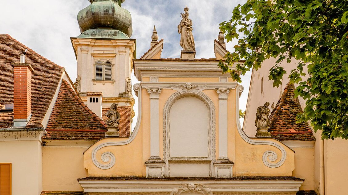 Ein historisches Tor mit einer eleganten Architektur und einer hübschen Turmspitze. Es ist von Bäumen umgeben und wirkt einladend. | © Wolfgang Speckner