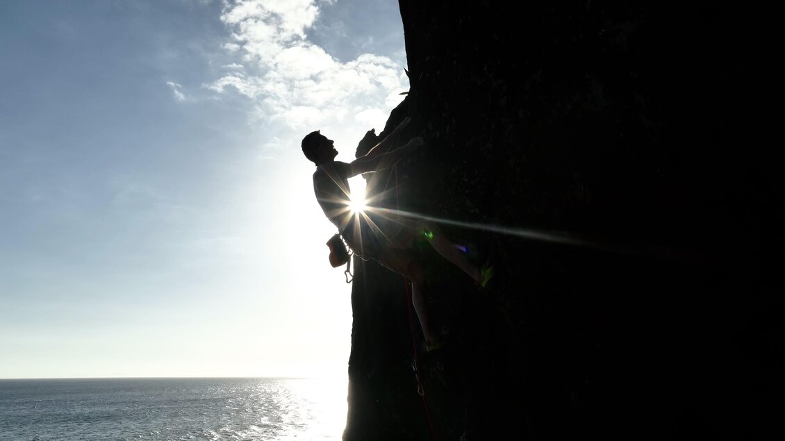 Ein Kletterer, der an einer Felswand hochsteigt, mit der Sonne im Hintergrund. Das Meer und der Himmel sind klar zu sehen. | © Benoit Regord