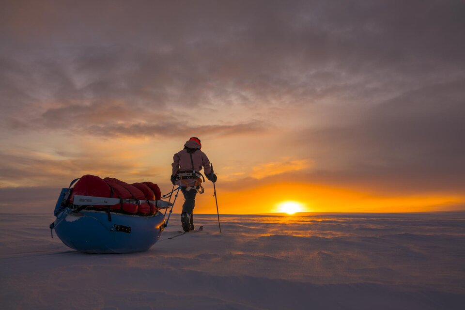Ein abenteuerlicher Mensch wandert durch eine schneebedeckte Landschaft, während die Sonne am Horizont aufgeht. Hinter ihm befindet sich ein roter Schlitten mit Gepäck. | © Caroline Cote + Vincent Colliard