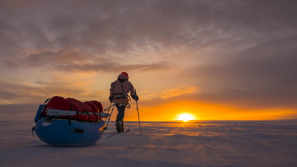 Ein abenteuerlicher Mensch wandert durch eine schneebedeckte Landschaft, während die Sonne am Horizont aufgeht. Hinter ihm befindet sich ein roter Schlitten mit Gepäck. | © Caroline Cote + Vincent Colliard