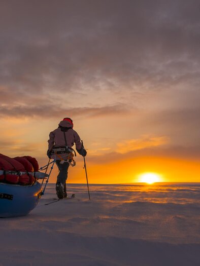 An adventurous person wanders through a snow-covered landscape as the sun rises on the horizon. Behind him is a red sled with luggage. | © Caroline Cote + Vincent Colliard