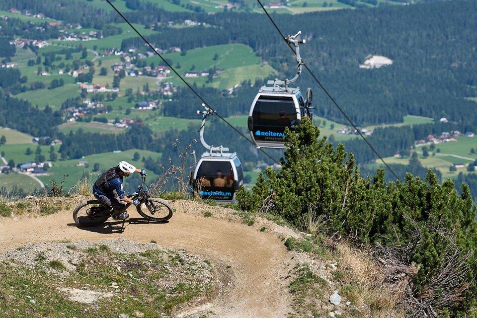 Ein Mountainbiker fährt auf einem schmalen Weg in den Bergen. Im Hintergrund sind Seilbahnen und eine malerische Landschaft zu sehen. | © Mr.EIT Reiteralm