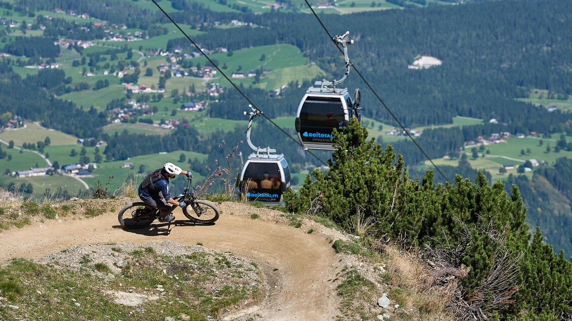 Ein Mountainbiker fährt auf einem schmalen Weg in den Bergen. Im Hintergrund sind Seilbahnen und eine malerische Landschaft zu sehen. | © Mr.EIT Reiteralm