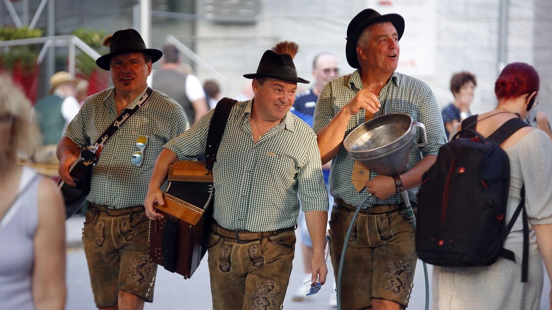 Drei Männer in traditioneller bayerischer Tracht gehen fröhlich die Straße entlang. Sie tragen Musikinstrumente und lächeln, während Passanten an ihnen vorbeigehen. | © Oststeiermark Tourismus
