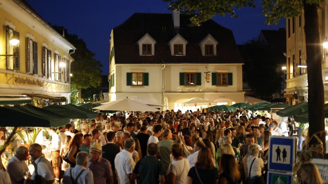 Eine belebte Straßenfeier bei Nacht mit vielen Menschen und festlicher Beleuchtung. Zahlreiche Tische und Sonnenschirme laden zum Verweilen ein. | © Oststeiermark Tourismus