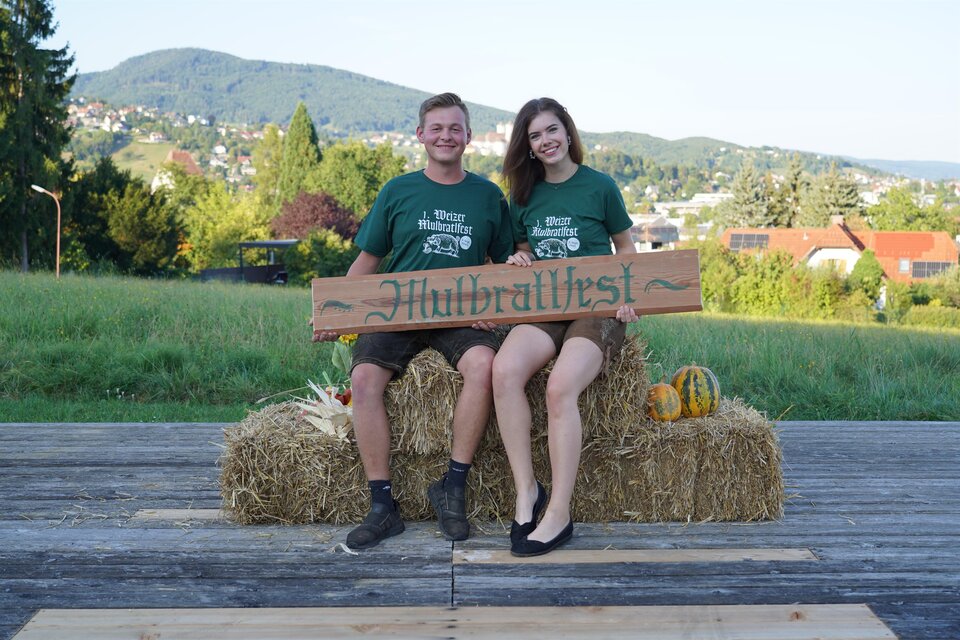 Zwei Personen sitzen auf Strohballen und halten ein Schild mit der Aufschrift "Hübraltfest". Im Hintergrund sind grüne Hügel und Häuser zu sehen. | © Oststeiermark Tourismus
