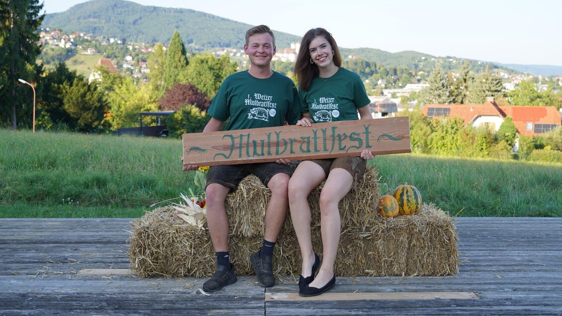 Zwei Personen sitzen auf Strohballen und halten ein Schild mit der Aufschrift "Hübraltfest". Im Hintergrund sind grüne Hügel und Häuser zu sehen. | © Oststeiermark Tourismus