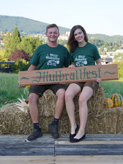 Zwei Personen sitzen auf Strohballen und halten ein Schild mit der Aufschrift "Hübraltfest". Im Hintergrund sind grüne Hügel und Häuser zu sehen. | © Oststeiermark Tourismus