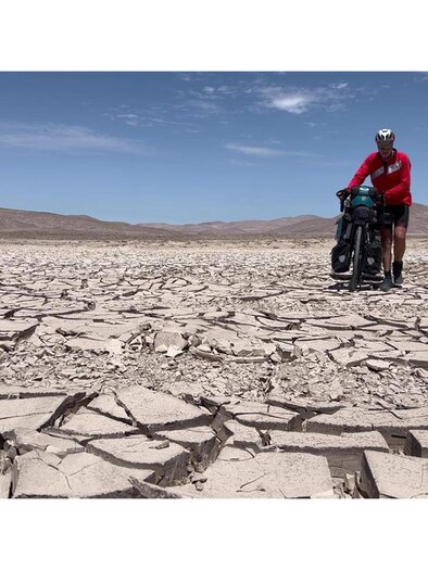 Radreisender in roter Jacke mit voll bepacktem Fahrrad auf ausgetrocknetem, rissigem Boden vor einer kargen Berglandschaft. | © Thomas Widerin