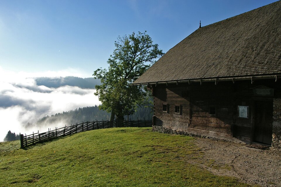 Eine alte Holzhütte steht auf einer grünen Wiese mit Blick auf neblige Täler. Im Hintergrund sind Bäume und der klare Himmel sichtbar. | © Jakob Hiller