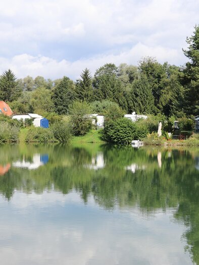 Ein ruhiger See mit Spiegelungen der umliegenden Bäume und Häuser. Im Hintergrund sind grüne Wiesen und ein blauer Himmel zu sehen. | © Erlebnisregion Murtal