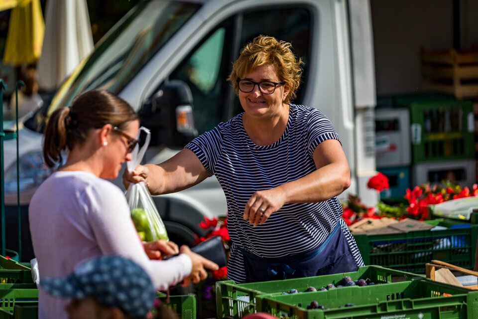 Eine Verkäuferin übergibt einer Kundin eine Tüte mit frischem Obst auf einem Markt. Im Hintergrund sind verschiedene Obstkisten und ein Lieferwagen zu sehen.