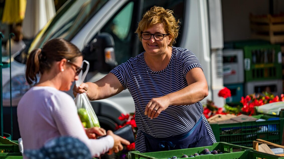Eine Verkäuferin übergibt einer Kundin eine Tüte mit frischem Obst auf einem Markt. Im Hintergrund sind verschiedene Obstkisten und ein Lieferwagen zu sehen.