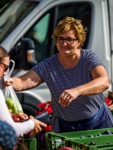Eine Verkäuferin übergibt einer Kundin eine Tüte mit frischem Obst auf einem Markt. Im Hintergrund sind verschiedene Obstkisten und ein Lieferwagen zu sehen.