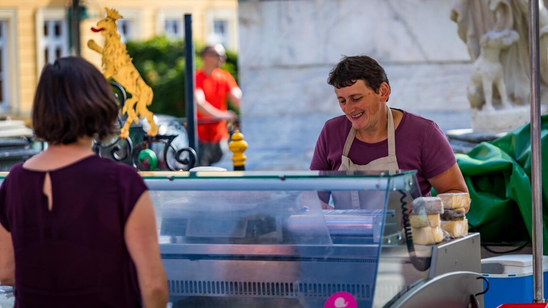 Eine Verkäuferin an einem Stand mit Eiscreme lächelt freundlich. Im Hintergrund sind weitere Menschen und eine Statue zu sehen.