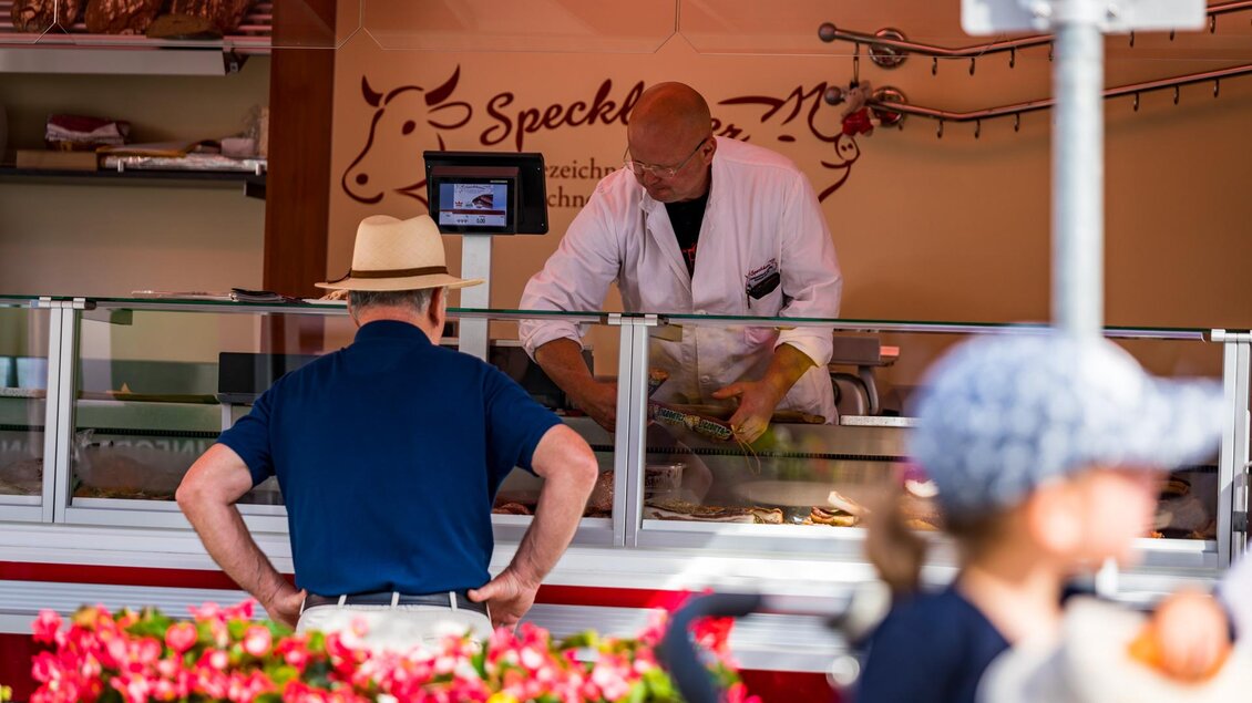 Ein Verkaufsstand mit einem Verkäufer, der an einem Wurststand arbeitet. Im Vordergrund steht ein Mann mit einem Hut und beobachtet die Aktion.