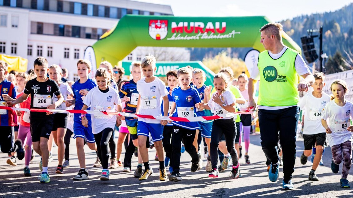 Eine Gruppe von Kindern läuft fröhlich bei einem Wettkampf. Im Hintergrund ist ein bunter Bogen mit dem Schriftzug „Murau“ zu sehen. | © Tv Murau