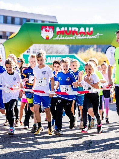 A group of children is happily running in a competition. In the background, a colorful arch with the inscription "Murau" can be seen. | © Tv Murau