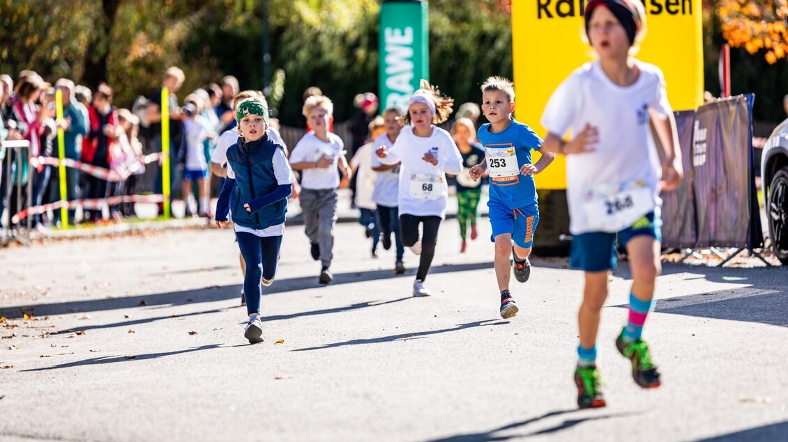 Eine Gruppe von Kindern läuft enthusiastisch in einem Wettlauf. Im Hintergrund stehen Zuschauer und es ist ein sonniger Tag. | © TV Murau