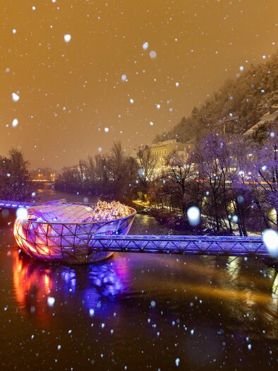 Un ponte innevato che attraversa un fiume è colorato da luci. Intorno ci sono alberi coperti di neve e un'atmosfera invernale delicata. | © (c) Graz Tourismus - Harry Schiffer