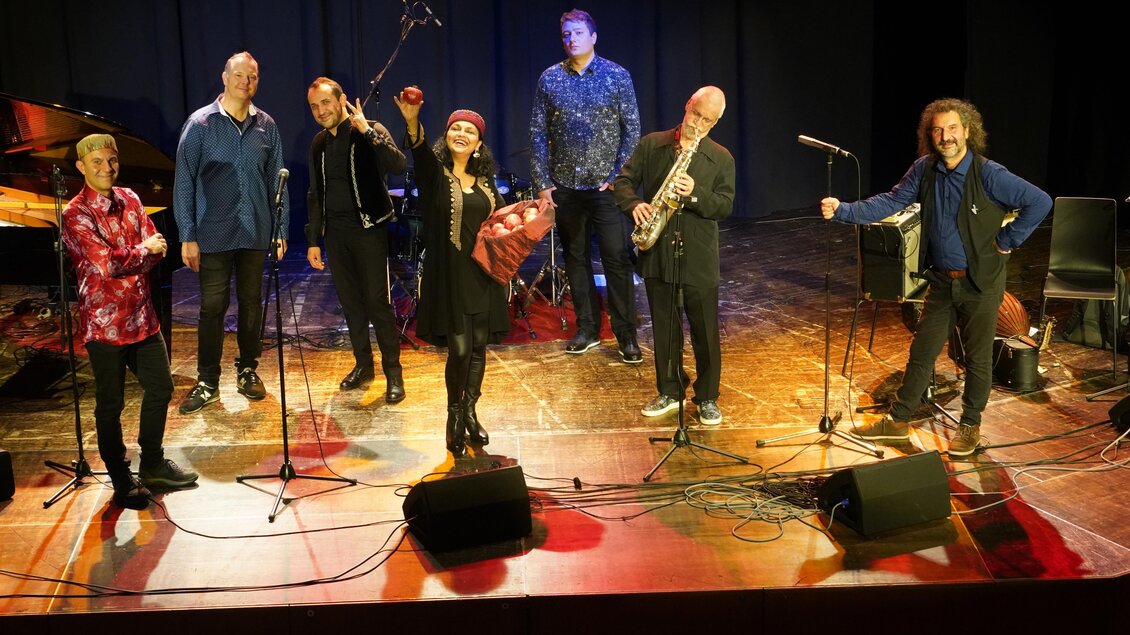 Eine Gruppe von Musikern steht auf einer Bühne mit Mikrofonen. Die Stimmung ist festlich und die Musiker lächeln in die Kamera. | © (c) Manfred Fiedler.