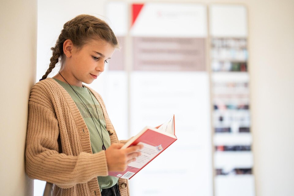 Ein Mädchen steht an einer Wand und liest ein Buch. Sie trägt einen gemütlichen Pullover und hat einen nachdenklichen Gesichtsausdruck. | © Stift Admont