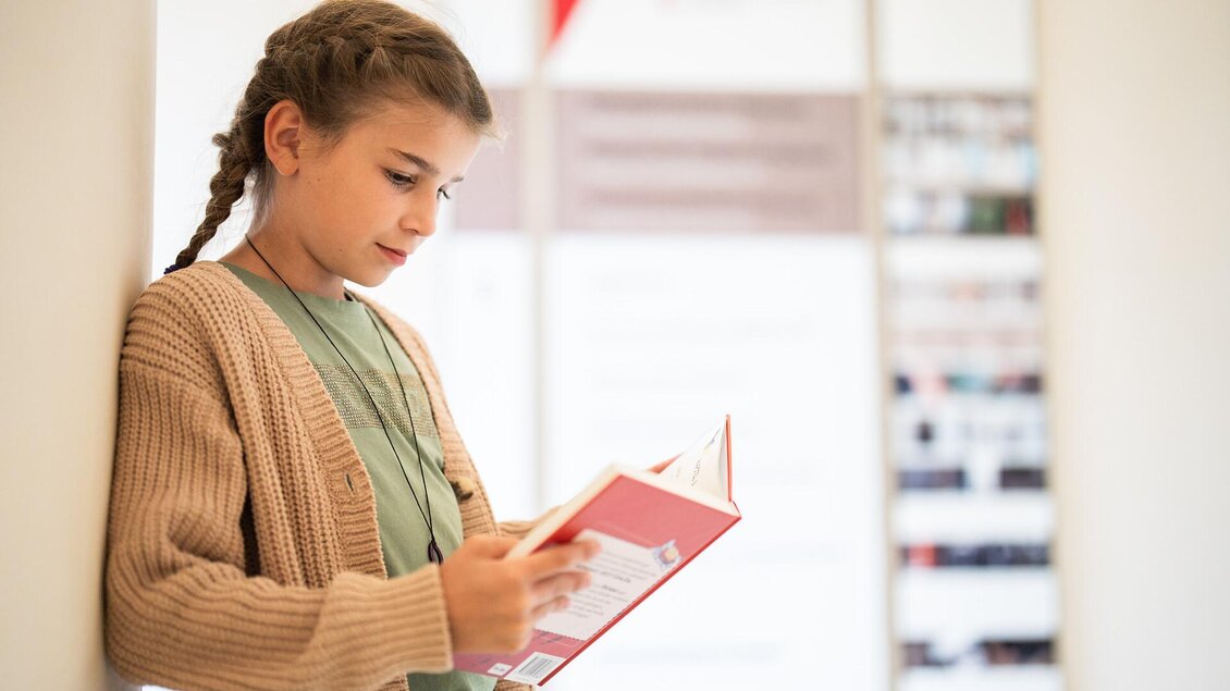 Ein Mädchen steht an einer Wand und liest ein Buch. Sie trägt einen gemütlichen Pullover und hat einen nachdenklichen Gesichtsausdruck. | © Stift Admont