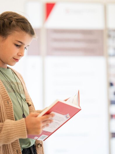 Ein Mädchen steht an einer Wand und liest ein Buch. Sie trägt einen gemütlichen Pullover und hat einen nachdenklichen Gesichtsausdruck. | © Stift Admont