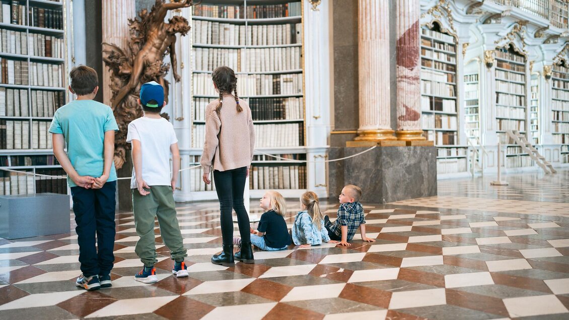 Eine Gruppe von Kindern steht vor einer beeindruckenden Bibliothek mit hohen Regalen voller Bücher. Der Boden ist mit einem schönen Schachbrettmuster gestaltet. | © Stift Admont
