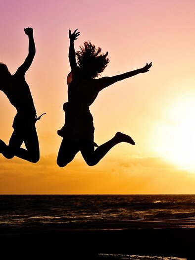 Two people are happily jumping into the sunset. The sky is painted in warm colors and the sea is visible in the background. | © Pixabay