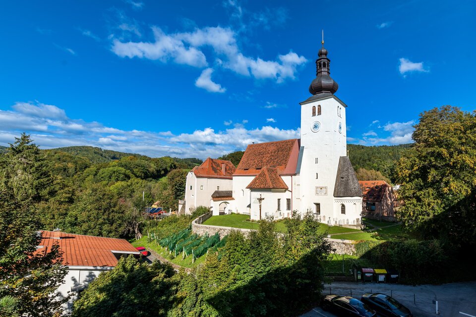 Eine charmante Kirche mit einem hohen Turm, umgeben von grünen Bäumen und blauen Himmel. Im Vordergrund sind einige Häuser und Parkplätze sichtbar. | © Die Abbilderei