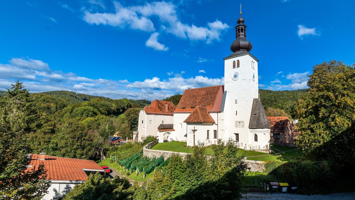 Eine charmante Kirche mit einem hohen Turm, umgeben von grünen Bäumen und blauen Himmel. Im Vordergrund sind einige Häuser und Parkplätze sichtbar. | © Die Abbilderei
