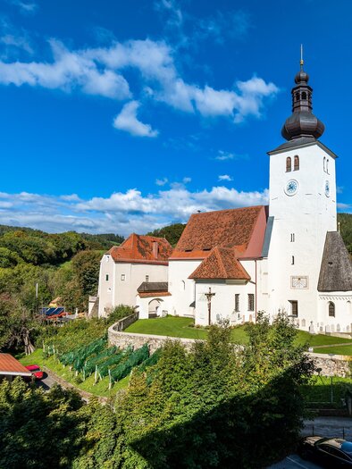 Eine charmante Kirche mit einem hohen Turm, umgeben von grünen Bäumen und blauen Himmel. Im Vordergrund sind einige Häuser und Parkplätze sichtbar. | © Die Abbilderei