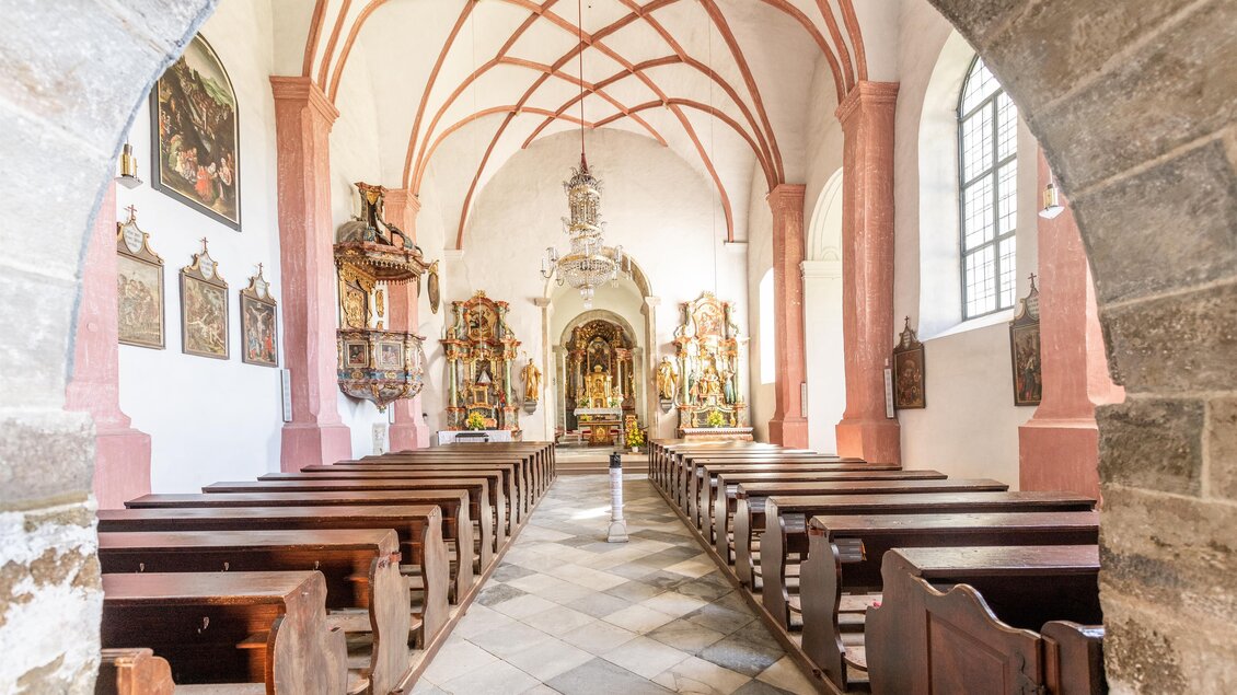 Eine Innenansicht einer Kirche mit Holzbänken und bunten Gemälden an den Wänden. Der Raum hat hohe, gewölbte Decken und wirkt lichtdurchflutet. | © Die Abbilderei