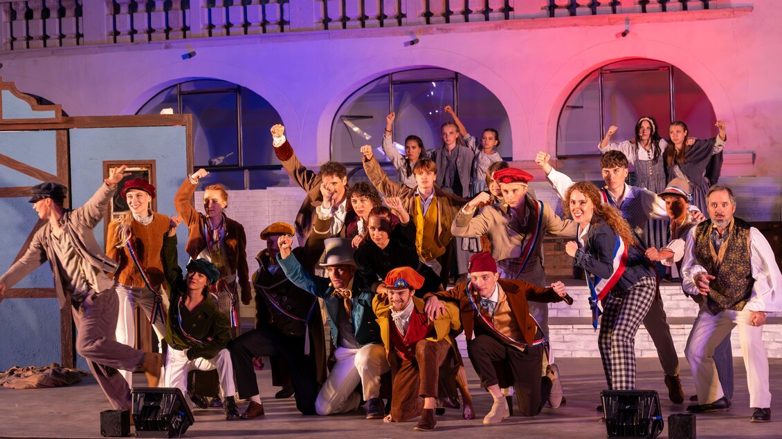 Eine lebhafte Theateraufführung mit einer Gruppe von Darstellern in bunten Kostümen. Die Schauspieler posieren fröhlich auf einer Bühne mit einem festlichen Hintergrund. | © Oststeiermark Tourismus