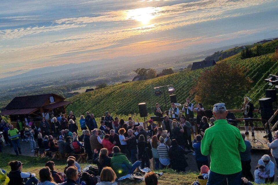 Eine Menschenmenge sitzt auf einer Wiese und genießt einen Sonnenuntergang. Im Hintergrund sind Weinreben und ein kleines Gebäude zu sehen. | © Weinlandkapelle Klöch