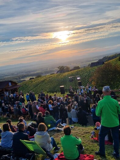 Eine Menschenmenge sitzt auf einer Wiese und genießt einen Sonnenuntergang. Im Hintergrund sind Weinreben und ein kleines Gebäude zu sehen. | © Weinlandkapelle Klöch
