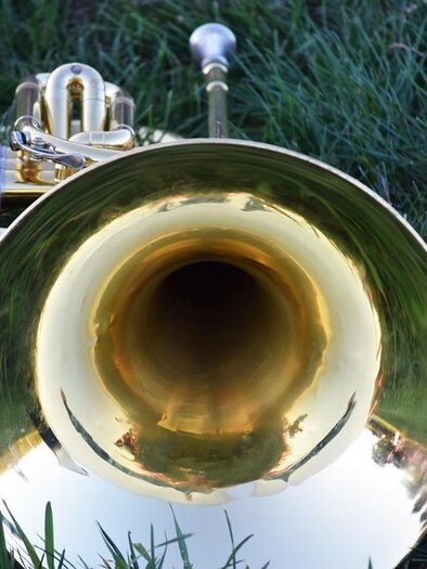 A large, shiny trumpet lies on green grass. The view inside the instrument shows the reflective surface. | © Pixabay