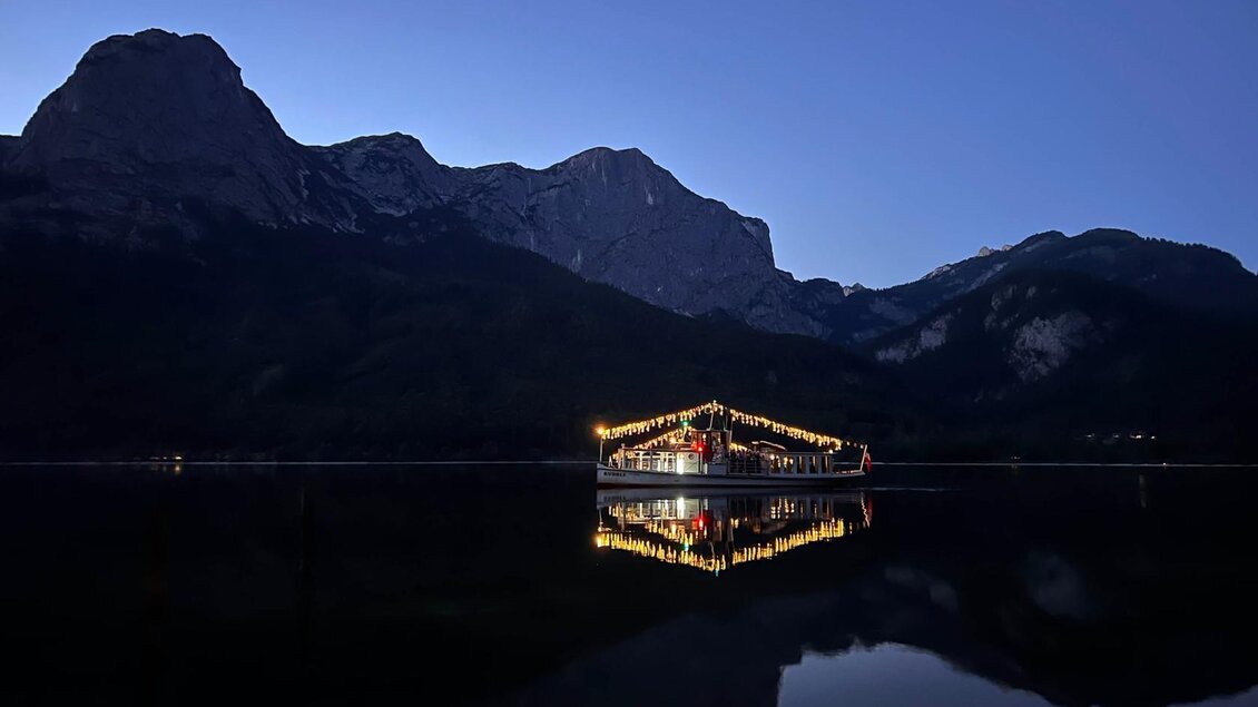 Ein ruhiger See mit einem beleuchteten Boot und majestätischen Bergen im Hintergrund. Der Himmel ist tiefblau und der Moment strahlt eine friedliche Stimmung aus. | © Schifffahrt Grundlsee Toplitzsee