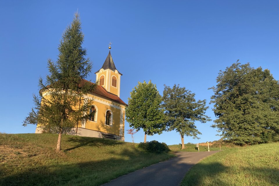 Idyllische Kapelle in Unterlamm, eingebettet in sanfte Hügel und umgeben von Bäumen – stimmungsvolle Landschaft bei warmem Abendlicht. | © Kapellengemeinschaft Haselbach