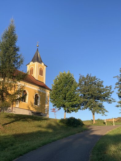 Idyllische Kapelle in Unterlamm, eingebettet in sanfte Hügel und umgeben von Bäumen – stimmungsvolle Landschaft bei warmem Abendlicht. | © Kapellengemeinschaft Haselbach