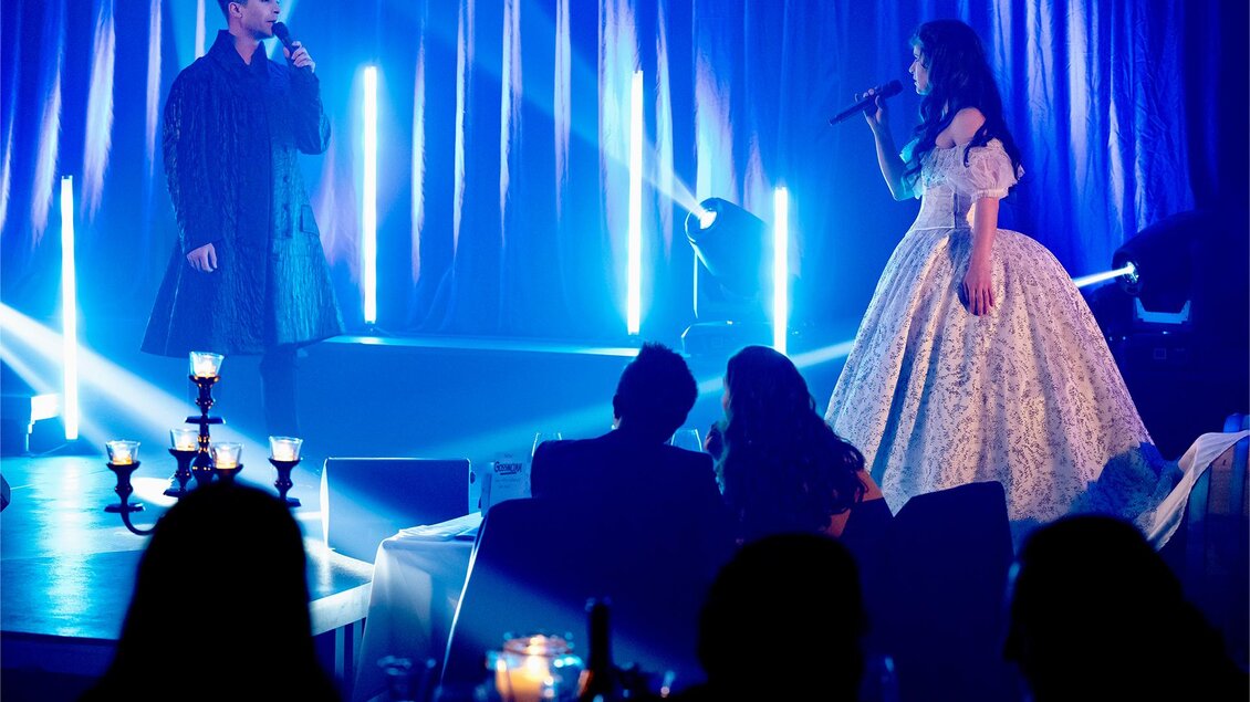 Ein musikalisches Duett auf einer Bühne. Zwei Sänger stehen in stilvollen Outfits vor beleuchtetem Hintergrund und performen für ein Publikum. | © Anja Wendzel