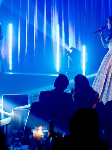 A musical duet on a stage. Two singers stand in stylish outfits in front of a lit background and perform for an audience. | © Anja Wendzel