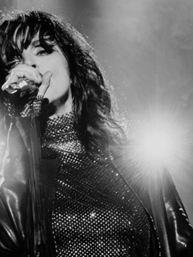 A black-and-white image of the singer NENA on stage with a microphone in hand. The lighting in the background creates a dramatic atmosphere. | © Sarah Rechbauer