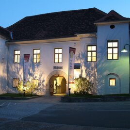Ein historisches Gebäude bei Nacht, beleuchtet mit warmem Licht. Die Fassade ist weiß mit großen Fenstern und einem einladenden Eingang.